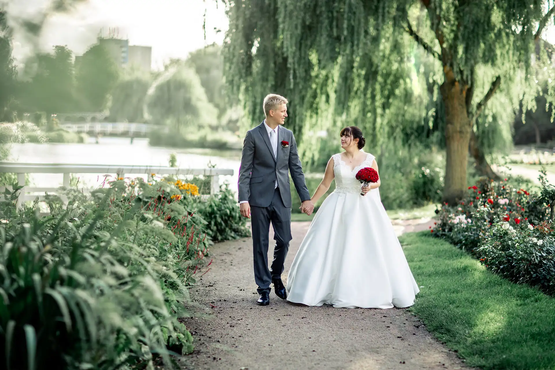Braut und Bräutigam gehen Hand in Hand einen Gartenweg entlang eines Sees und halten ihren besonderen Tag mit einem perfekten Foto fest. Die Braut trägt ein weißes Kleid und hält rote Blumen in der Hand, während der Bräutigam einen grauen Anzug mit einer roten Ansteckblume trägt. Üppiges Grün und eine Weide rahmen die Szene ein.