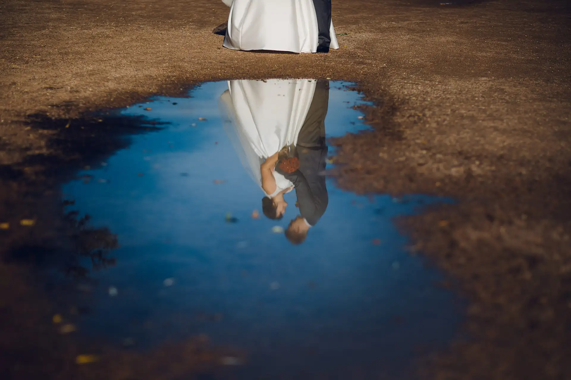 Bei einem bezaubernden Hochzeitsshooting spiegelt sich ein Brautpaar wunderschön in einer Pfütze auf einem Feldweg. Das weiße Kleid der Braut und der dunkle Anzug des Bräutigams verschmelzen unter dem klaren blauen Himmel zu einer Umarmung.