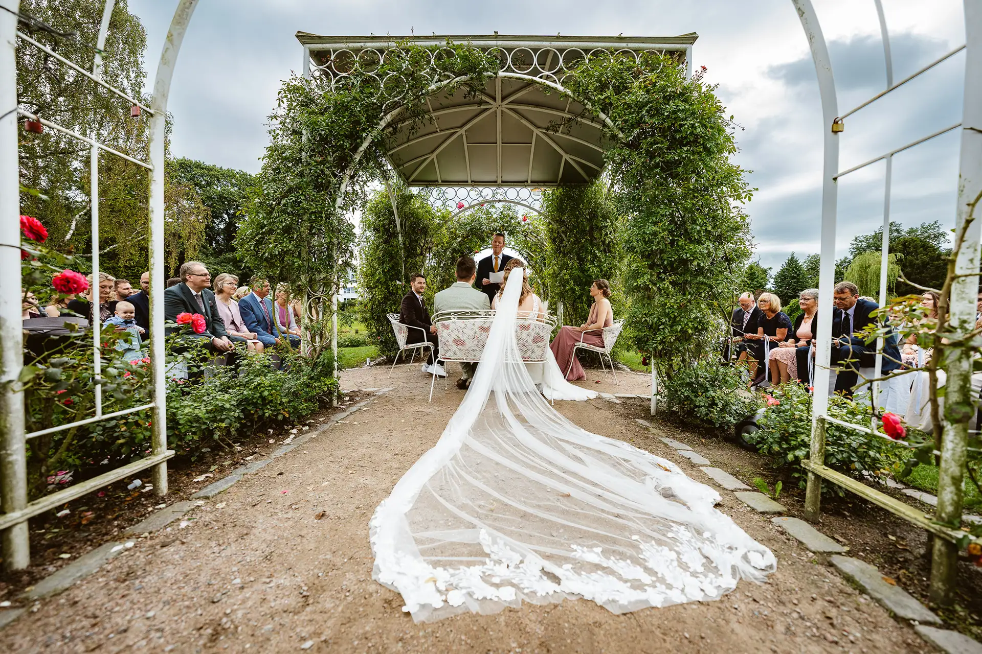 Rosarium Uetersen, Standesbeamter steht auf der Hochzeitsinsel im Rosarium und redet zum Brautpaar. Langer weißer Schleier fällt nach hinten auf den Boden.